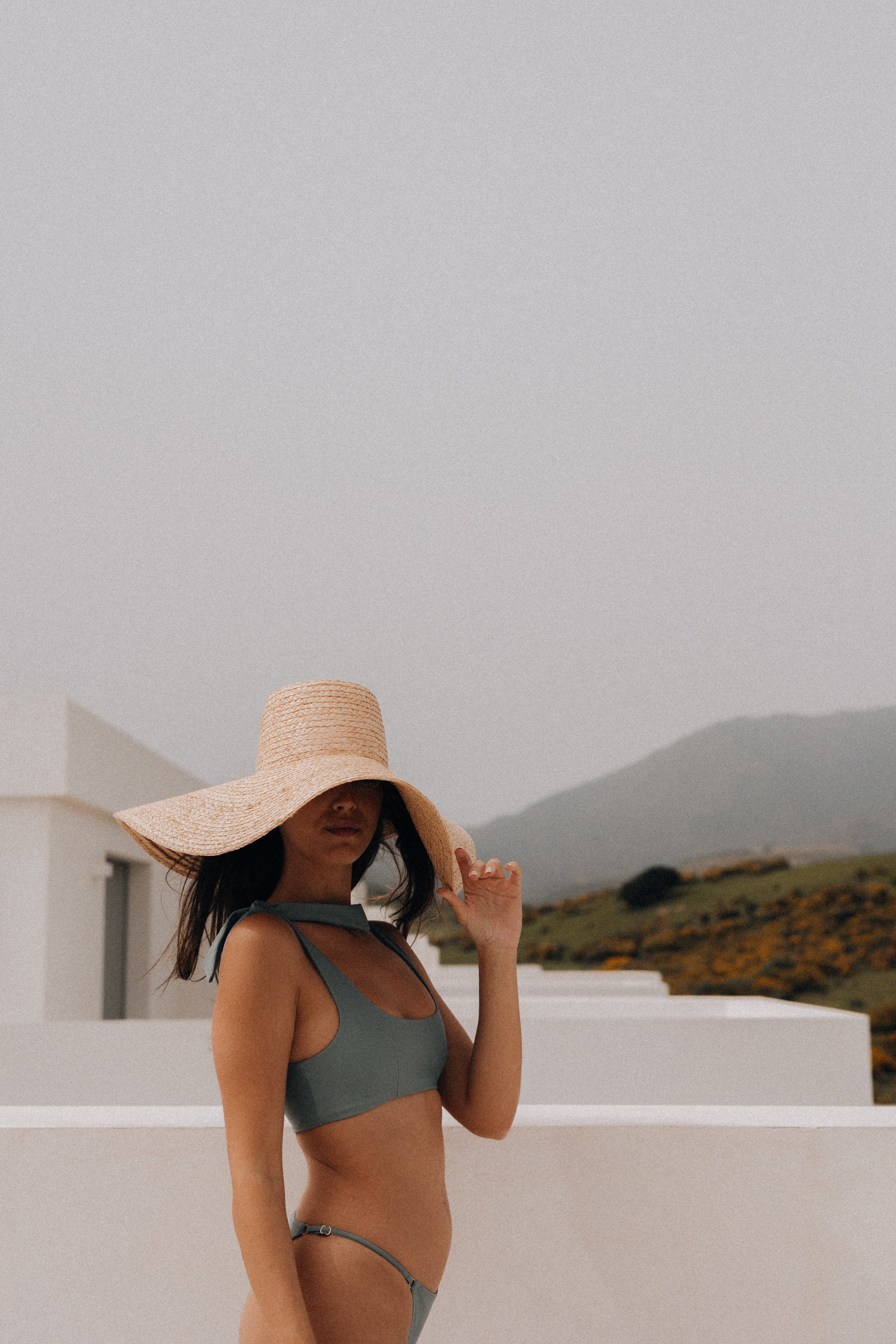 Woman wearing a wide-brimmed hat and green bikini against a mountainous landscape.